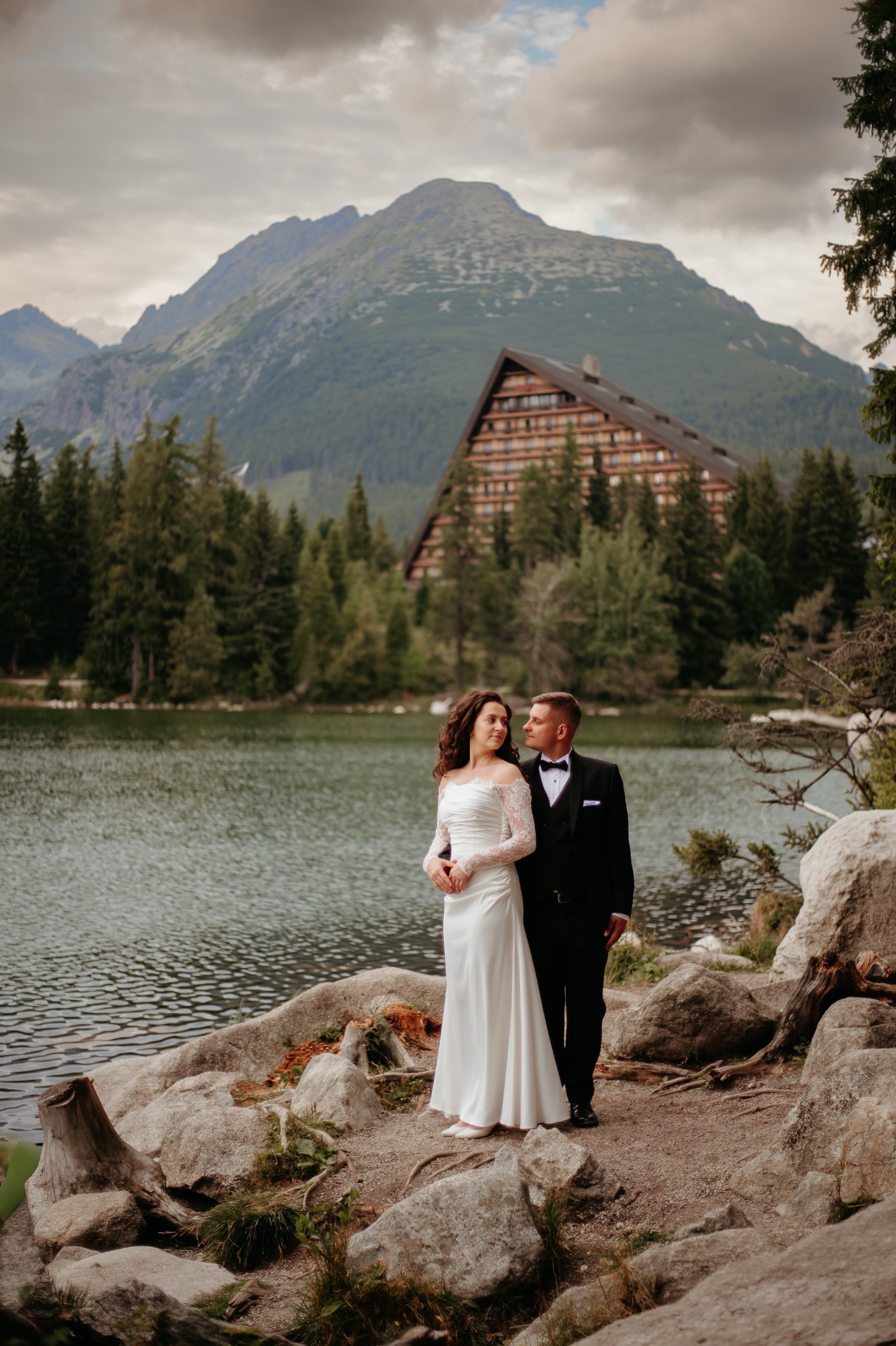 Para młoda w eleganckich strojach pozuje nad jeziorem Morskie Oko, z widokiem na góry i budynek w tle. Romantyczna sceneria, naturalne światło, profesjonalna fotografia.