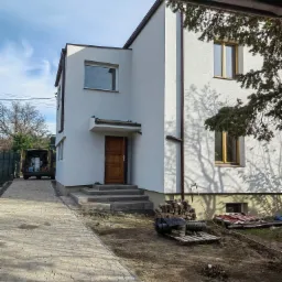 Nowoczesny, dwukondygnacyjny dom z białą elewacją i drewnianymi oknami, widok z boku na świeżo wykończony budynek z podjazdem z kostki brukowej i zaparkowanym vanem z otwartymi drzwiami...