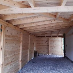 Usługi Stolarskie Krzysztof Torczyński - Wnętrze drewnianej konstrukcji w trakcie budowy, widoczne surowe deski ścian i dachu, podłoże wysypane żwirem, surowy stan wykończenia.