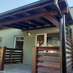 Zadaszona, drewniana konstrukcja tarasowa z ciemnym orynnowaniem, przylegająca do budynku o zielonej elewacji, z drewnianą balustradą w tym samym odcieniu.