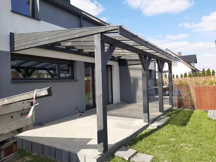 Zadaszenie tarasu z ciemnego drewna, wsparte na solidnych słupach, przylegające do szarego domu z dużymi oknami, widoczne fragmenty trawnika i drabina oparta o ścianę budynku.