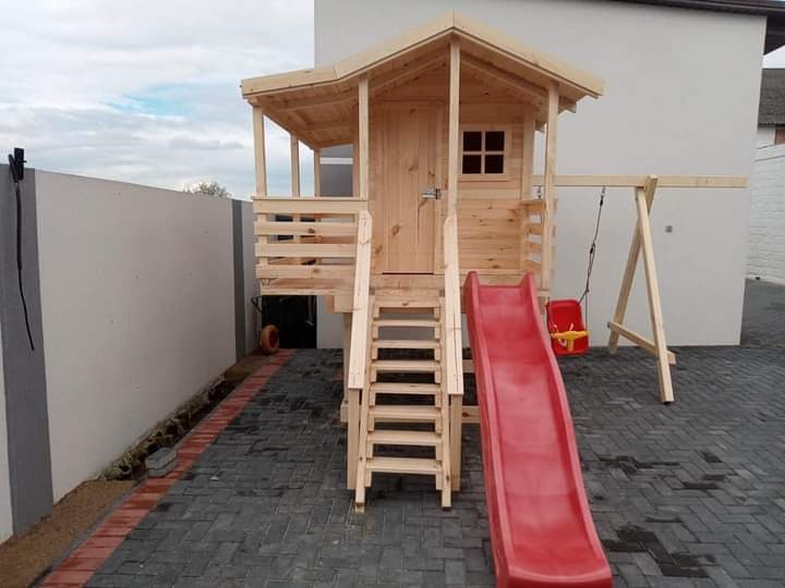 Drewniany plac zabaw dla dzieci z domkiem, schodami, zjeżdżalnią i huśtawką, ustawiony na brukowanym podwórku przy białym budynku i szarym ogrodzeniu.