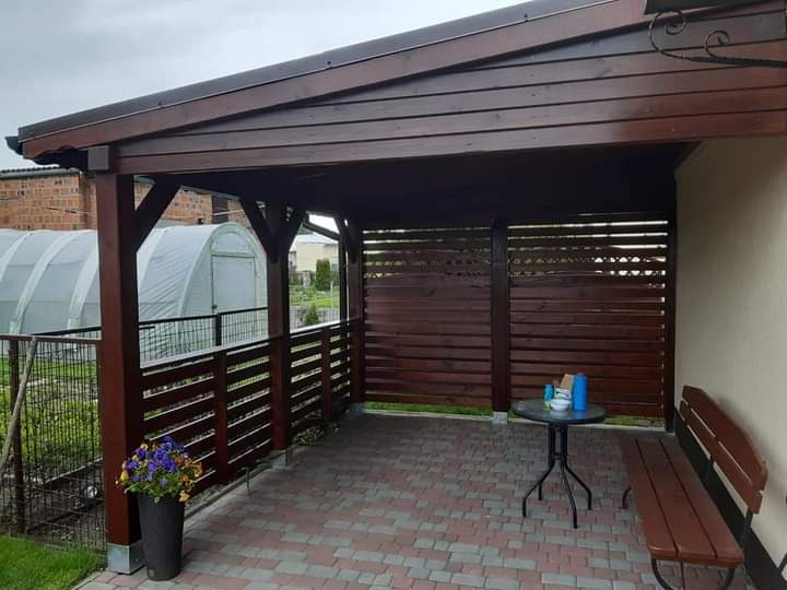 Zadaszona przestrzeń tarasowa z drewnianą konstrukcją, osłonięta ażurowymi panelami, z widocznym fragmentem ogrodu i szklarnią w tle.