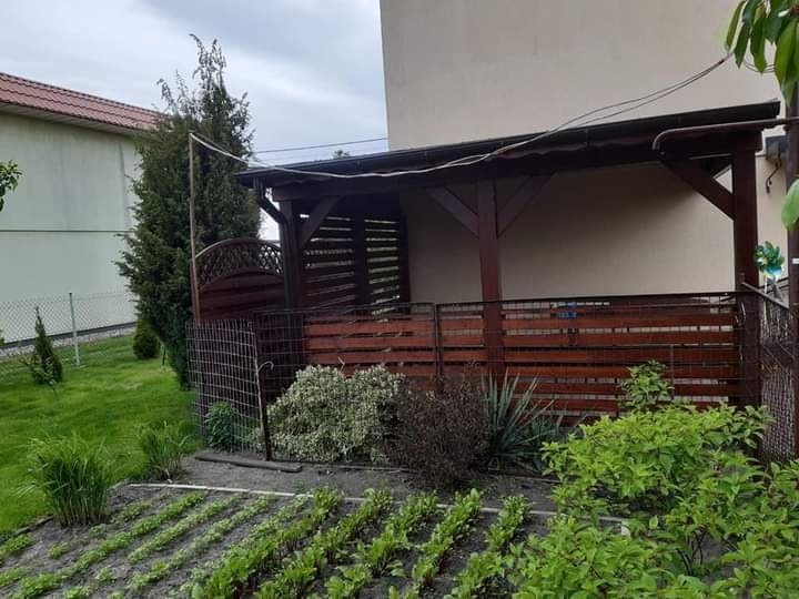 Zadaszona konstrukcja z ciemnego drewna z poziomymi deskami, przylegająca do budynku, widoczna z poziomu grządki warzywnej w ogrodzie.