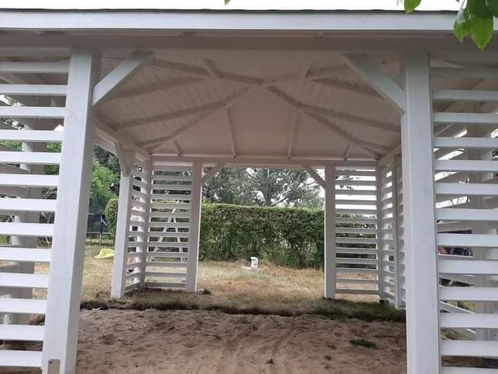 Biała, drewniana pergola z lamelami, widoczna z zewnątrz, z piaskiem na podłożu, otoczona zielenią.
