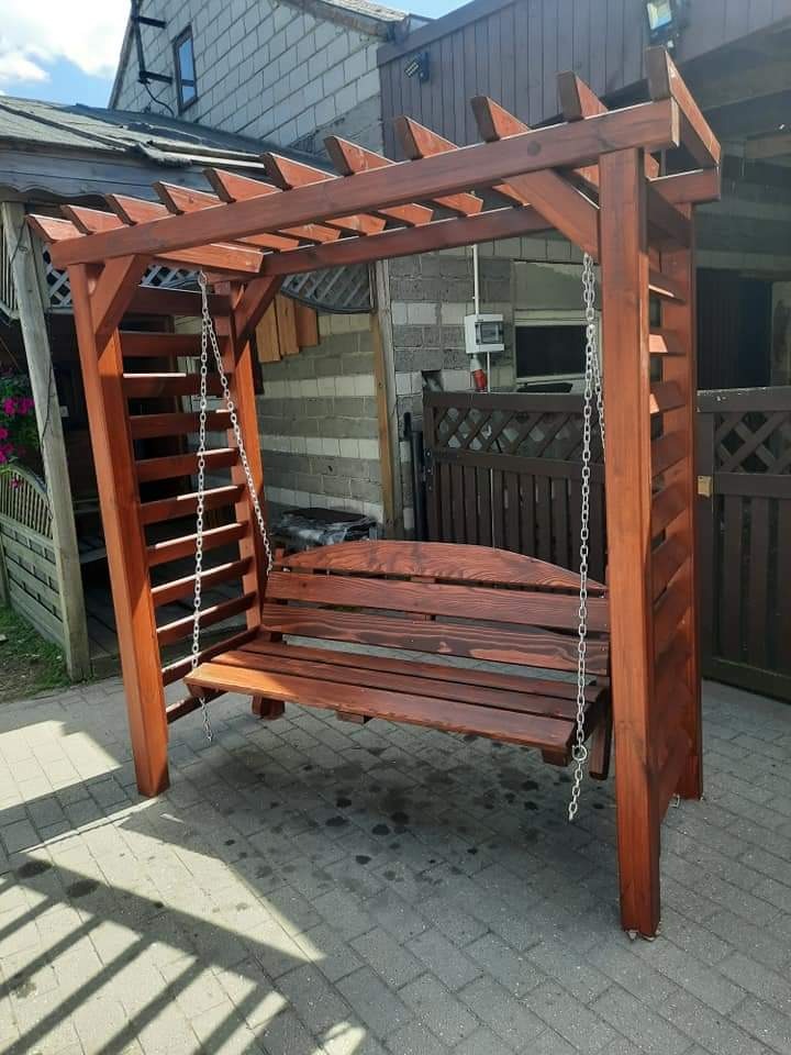 Drewniana huśtawka ogrodowa zadaszona pergolą, zawieszona na metalowych łańcuchach, usytuowana na brukowanym patio przed budynkiem z szarej cegły.