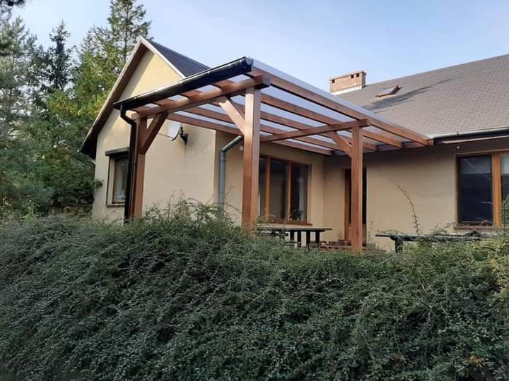 Drewniana konstrukcja z przezroczystym zadaszeniem przylegająca do budynku, otoczona zielenią.
