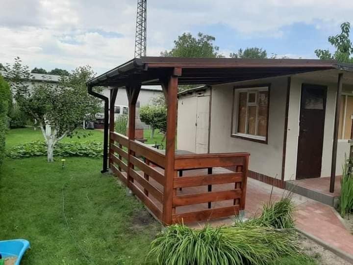 Zadaszone miejsce do siedzenia z drewnianą balustradą przy budynku gospodarczym, widok na trawnik i drzewa.