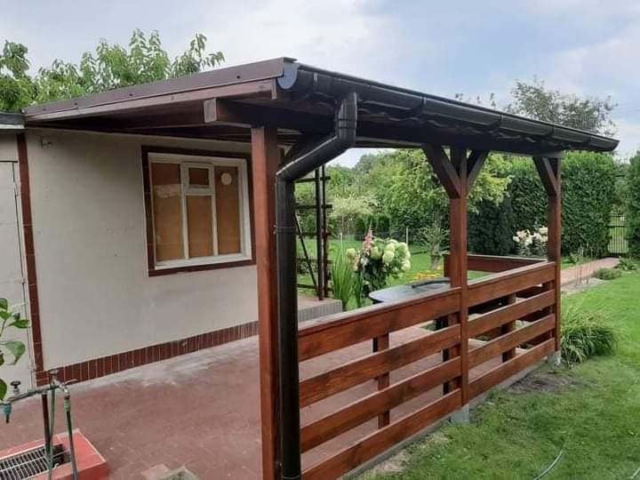 Drewniana konstrukcja z zadaszeniem, pełniącą funkcję zadaszonego tarasu, przylegająca do budynku z oknem, z widocznym systemem rynnowym i ogrodzeniem z poziomych desek, otoczona zielenią.