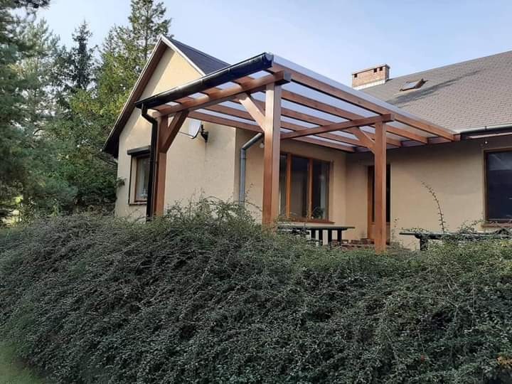 Drewniana konstrukcja z przezroczystym zadaszeniem, prawdopodobnie pergola, przylegająca do ściany domu. Widoczne drewniane słupy i belki oraz bujna zieleń krzewów na pierwszym planie.