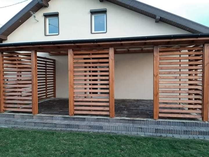 Drewniana pergola z poziomymi listwami, przylegająca do domu z dwoma oknami, wykończona kamiennymi schodami i trawnikiem.