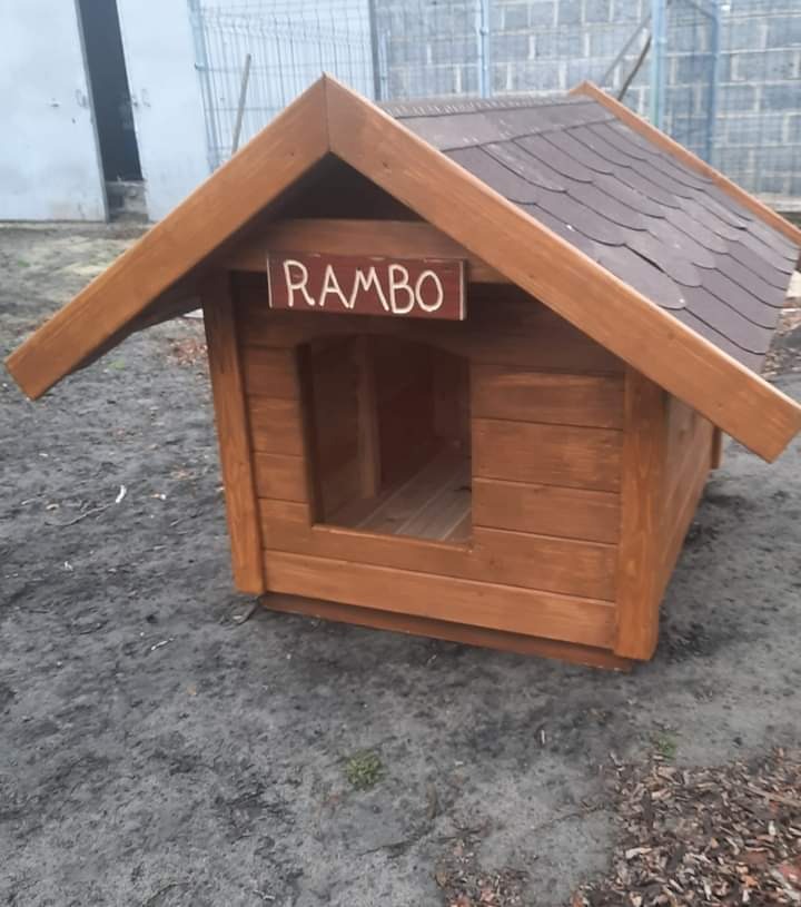 Drewniana buda dla psa z brązowym, spadzistym dachem i tabliczką z imieniem 'RAMBO', stojąca na ziemi.