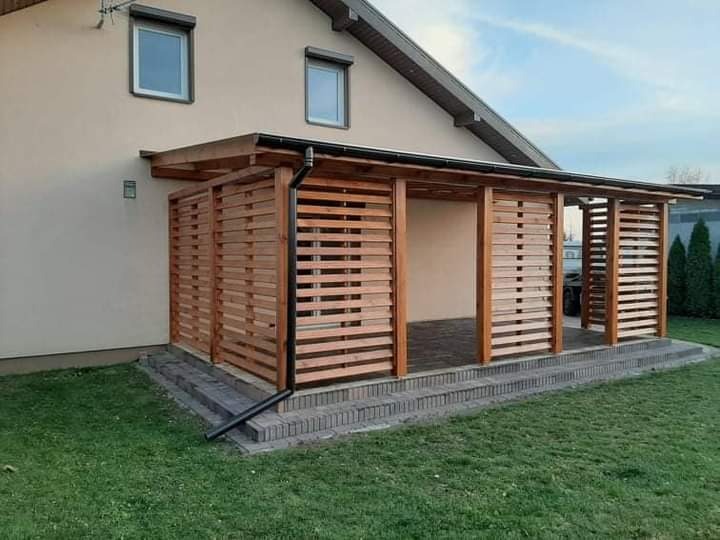 Zadaszona konstrukcja drewniana z poziomymi listwami, przylegająca do budynku mieszkalnego, z rynną odprowadzającą wodę i schodami z kostki brukowej.