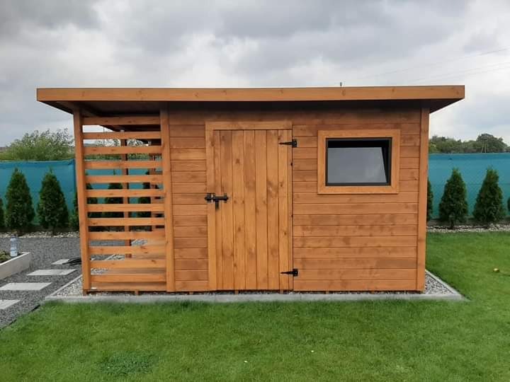 Drewniany domek narzędziowy z oknem i ażurową ścianką, wykończony w naturalnym odcieniu brązu, na tle zielonego trawnika i pochmurnego nieba.