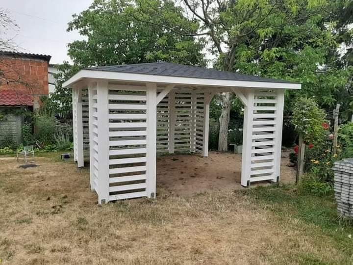 Biała, drewniana altana z czarnym dachem, wykonana z poziomych listew, stojąca na trawniku w ogrodzie. W tle widoczne drzewa i budynek z czerwonej cegły.