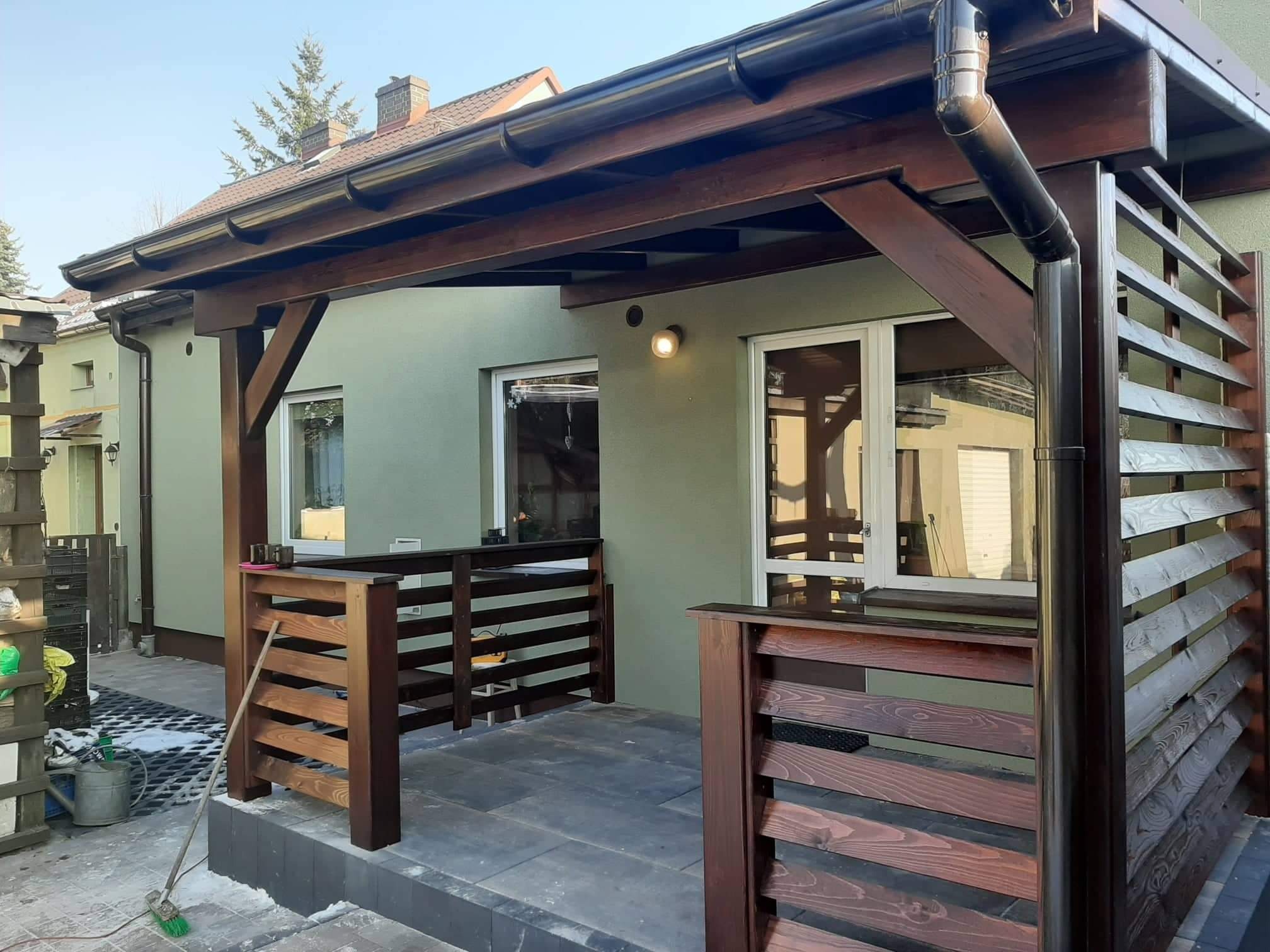 Zadaszony taras z drewnianą konstrukcją i poręczą, widoczne orynnowanie, ściany budynku w kolorze oliwkowym, zamiataczka i wiadro na posadzce.