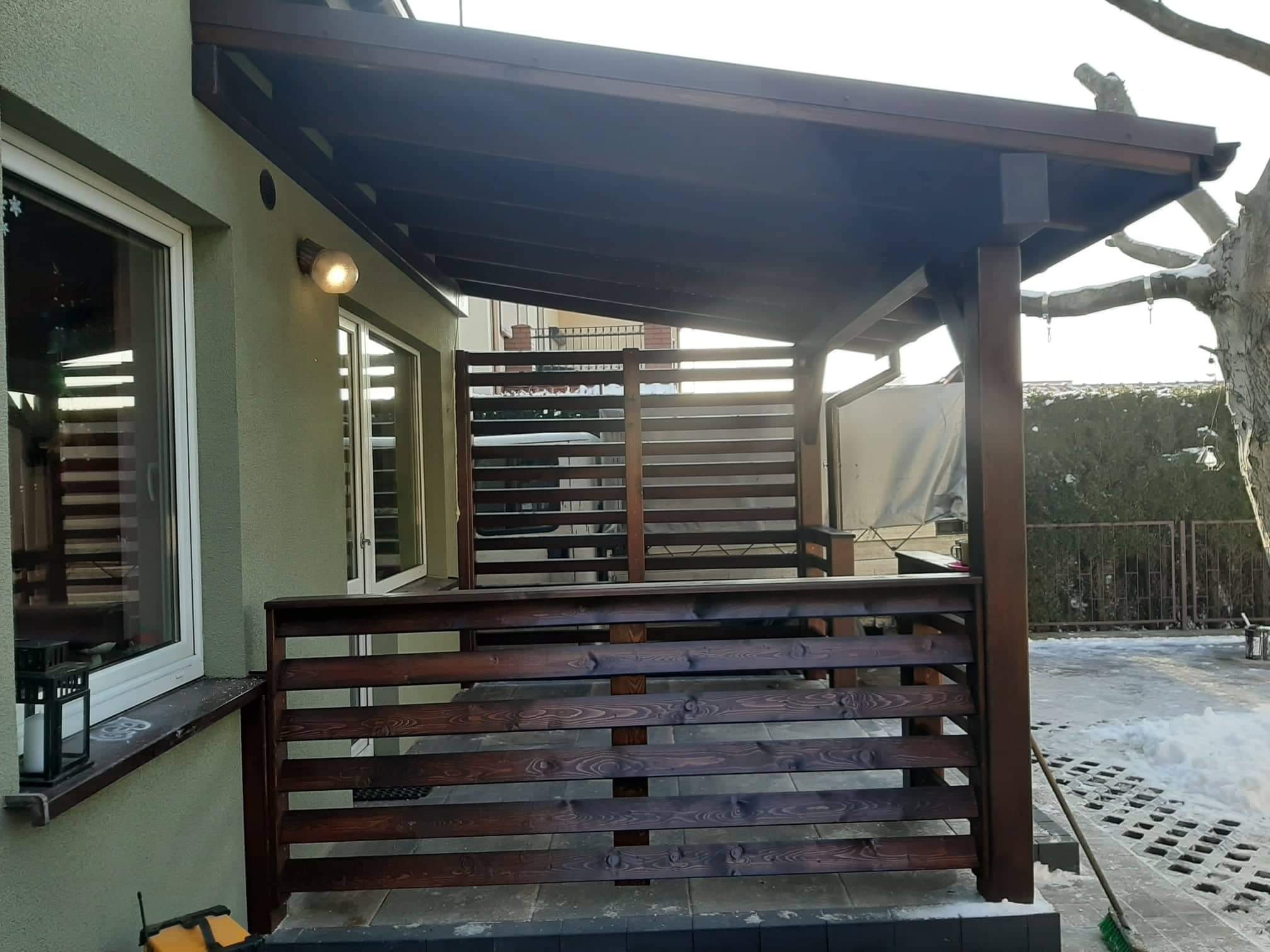 Zadaszony taras z drewnianą balustradą w zimowej scenerii, widok z boku domu z widoczną lampą zewnętrzną i fragmentem okna z dekoracją.