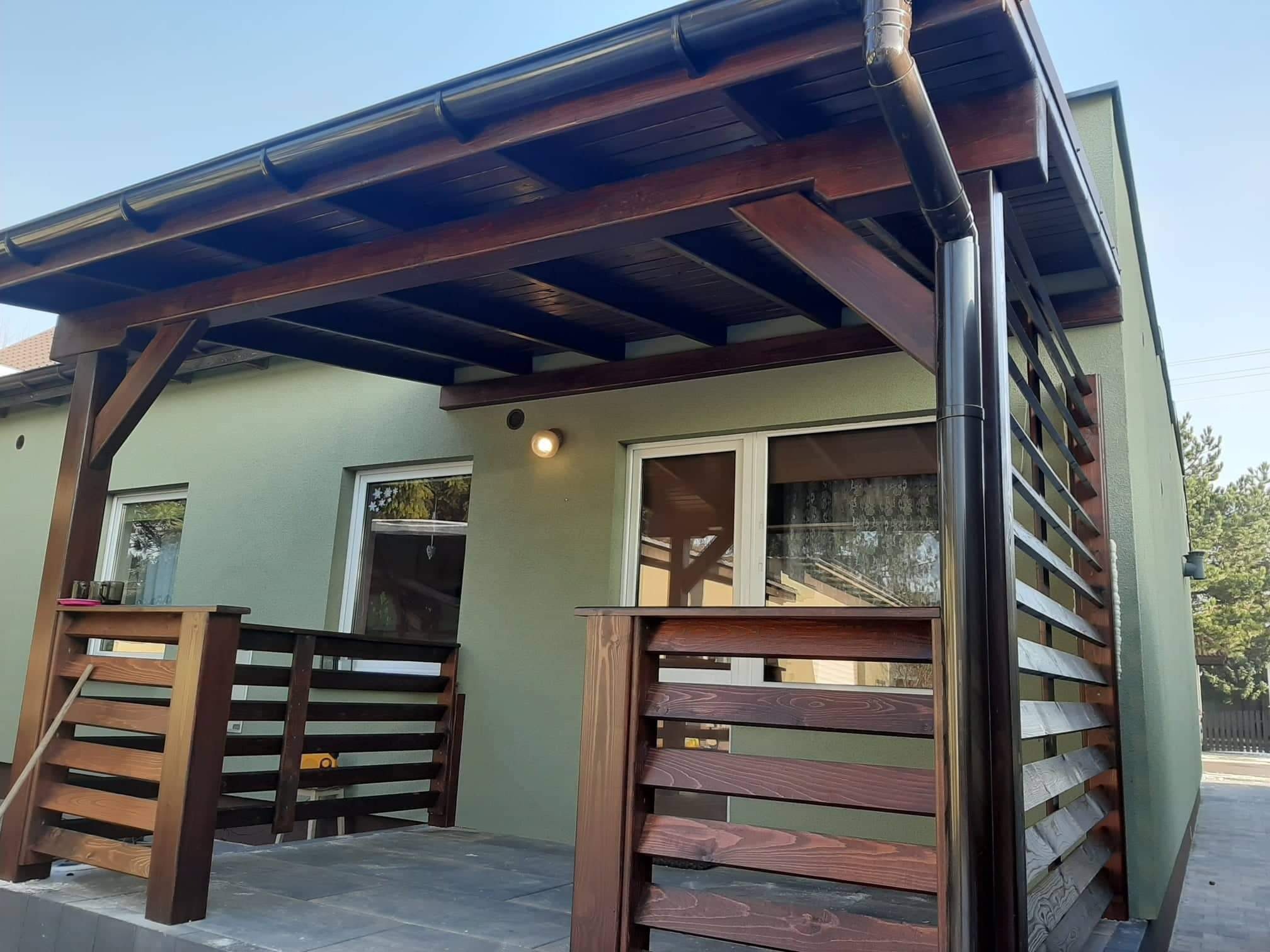Zadaszona, drewniana konstrukcja tarasowa z ciemnym orynnowaniem, przylegająca do budynku o zielonej elewacji, z drewnianą balustradą w tym samym odcieniu.