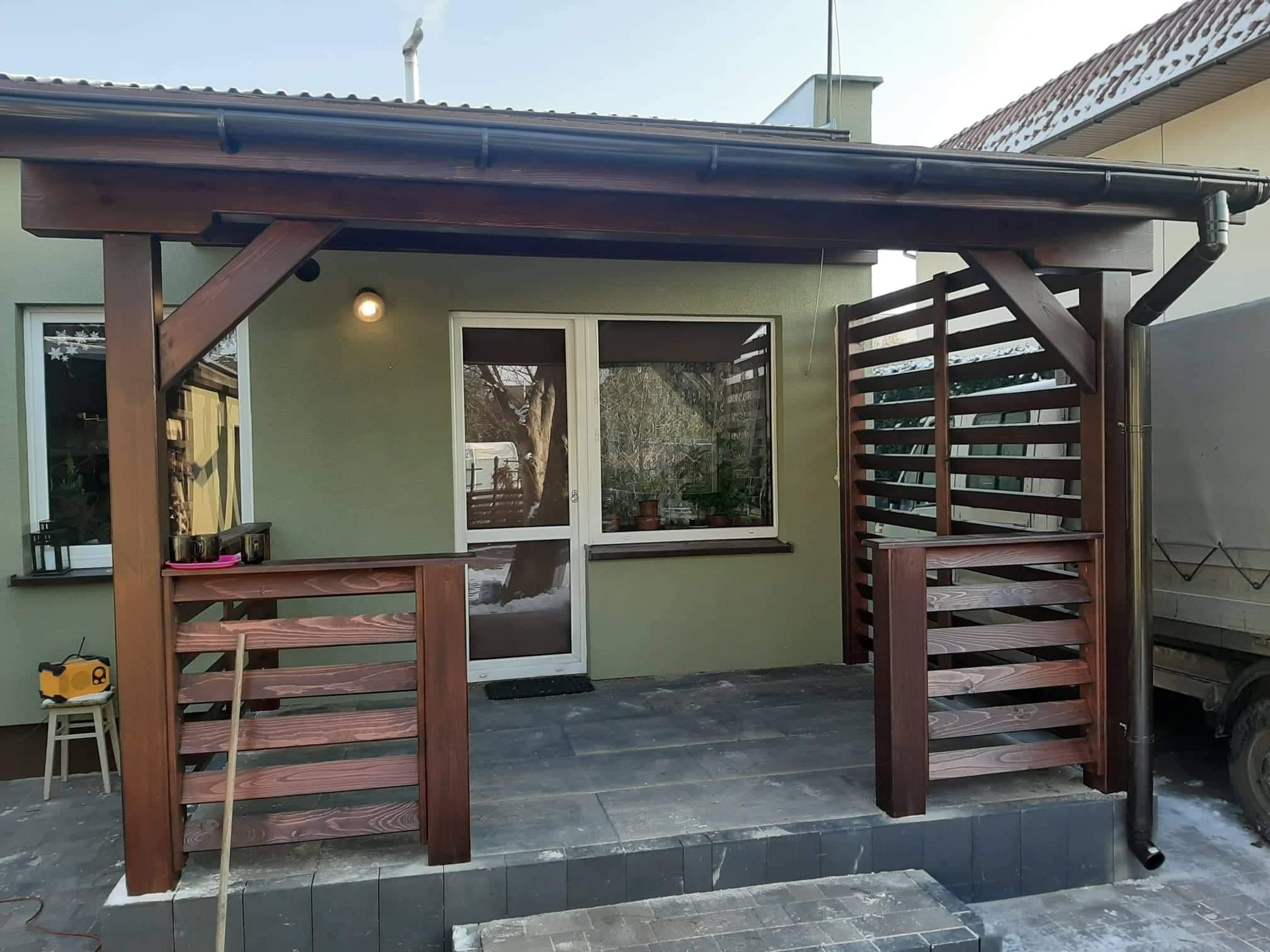 Zadaszony taras z drewnianą konstrukcją, wykończony w ciemnym brązie, z ażurową balustradą i wejściem z dwuskrzydłowymi drzwiami, na zewnątrz budynku z zieloną elewacją.