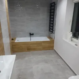 Wykończona łazienka z szarymi płytkami, drewnianą obudową wanny, czarną armaturą i grzejnikiem drabinkowym, widok na umywalkę i toaletę.