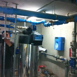 Sanitherm Project Sadowski Andrzej - Instalacja uzdatniania wody w kotłowni, widoczne rury w kolorze niebieskim, zbiorniki ze stali nierdzewnej, pracownik w trakcie montażu i kartony z elementami instalacji.