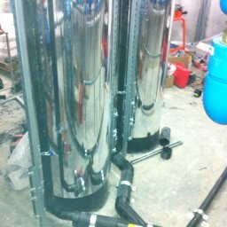 Sanitherm Project Sadowski Andrzej - Dwa chromowane zbiorniki z podłączonymi rurami kanalizacyjnymi, zamocowane do podłoża za pomocą metalowych wsporników; widoczny fragment wiertarki akumulatorowej.