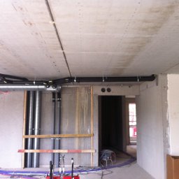 Sanitherm Project Sadowski Andrzej - Surowy betonowy strop z zamontowaną instalacją wentylacyjną i rurami kanalizacyjnymi, widoczne kable elektryczne na podłodze, w tle otwór drzwiowy i okno.