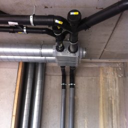 Sanitherm Project Sadowski Andrzej - Widok od dołu na skomplikowaną instalację rur wentylacyjnych i hydraulicznych z czarnymi i srebrnymi rurami pod betonowym sufitem. Widoczne złącza, obejmy i filtry.