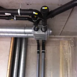 Widok od dołu na skomplikowaną instalację rur wentylacyjnych i hydraulicznych z czarnymi i srebrnymi rurami pod betonowym sufitem. Widoczne złącza, obejmy i filtry.