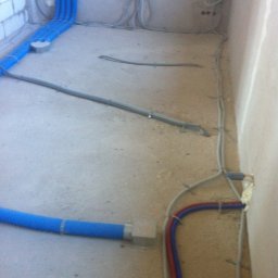 Sanitherm Project Sadowski Andrzej - Wnętrze w stanie surowym w Białymstoku z widocznymi elementami instalacji: niebieskimi rurami wentylacyjnymi, przewodami elektrycznymi i rurami grzewczymi w kolorach czerwonym i niebieskim...