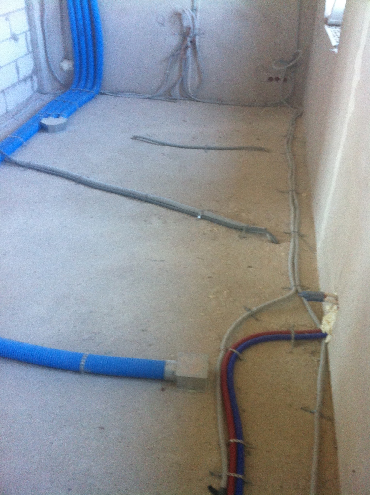 Wnętrze w stanie surowym w Białymstoku z widocznymi elementami instalacji: niebieskimi rurami wentylacyjnymi, przewodami elektrycznymi i rurami grzewczymi w kolorach czerwonym i niebieskim...