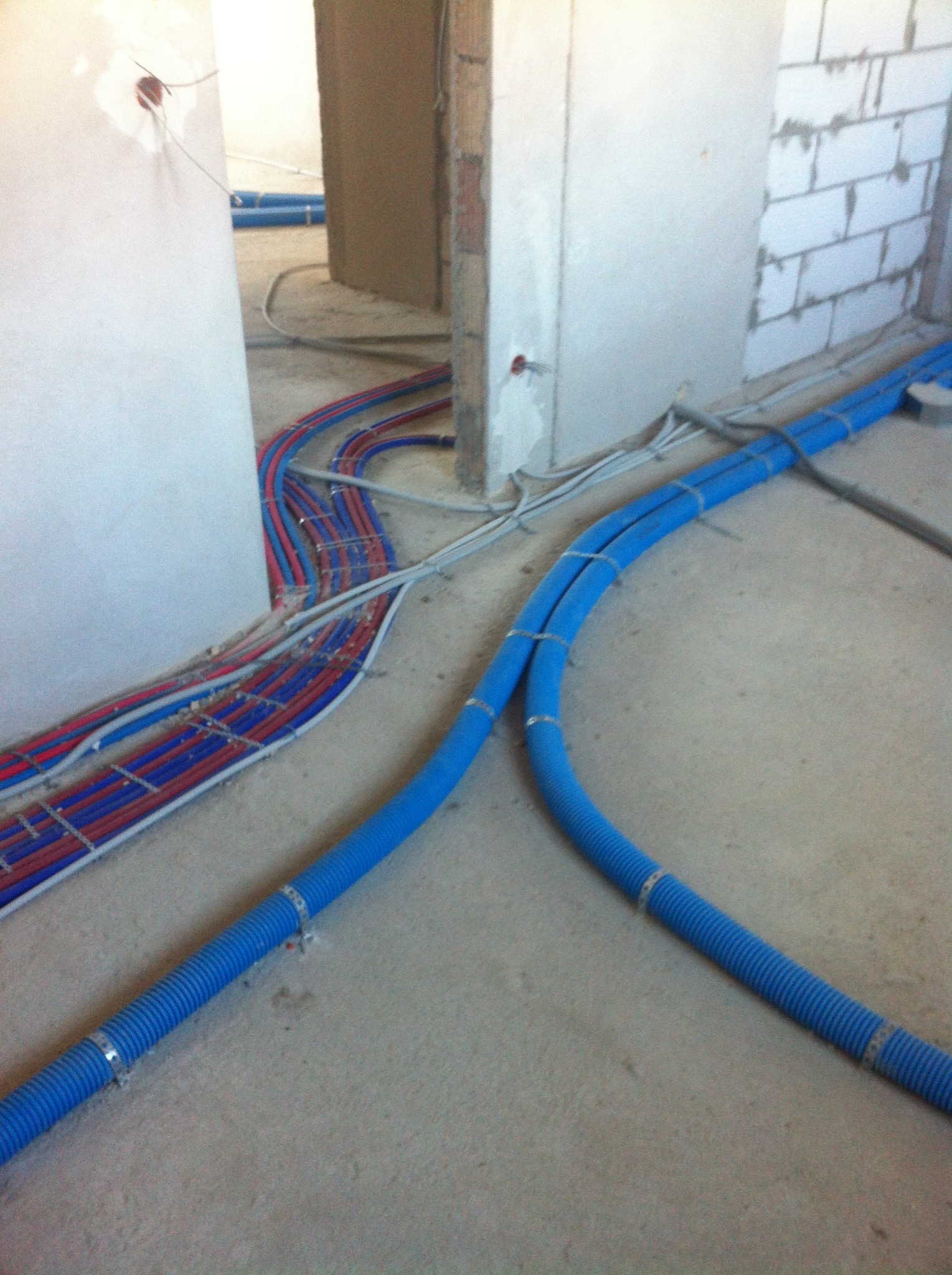 Węże grzewcze w niebieskiej karbowanej osłonie i czerwono-niebieskie rurki mocowane do podłoża w surowym stanie, widoczne ściany z betonu komórkowego i instalacje elektryczne.