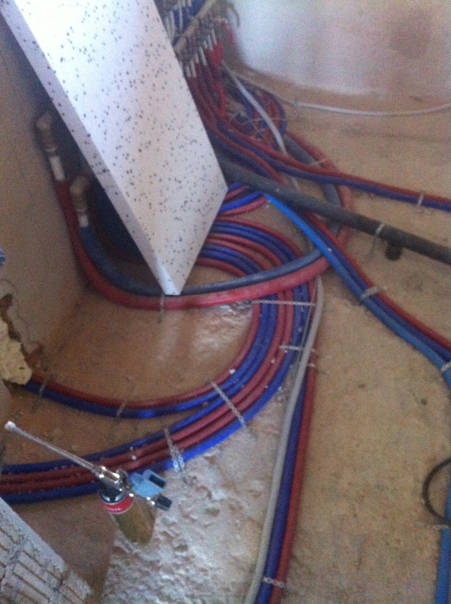 Wiązka czerwono-niebieskich rur instalacji podłogowej na surowym podłożu, z widocznym narzędziem do łączenia rur i fragmentem płyty.