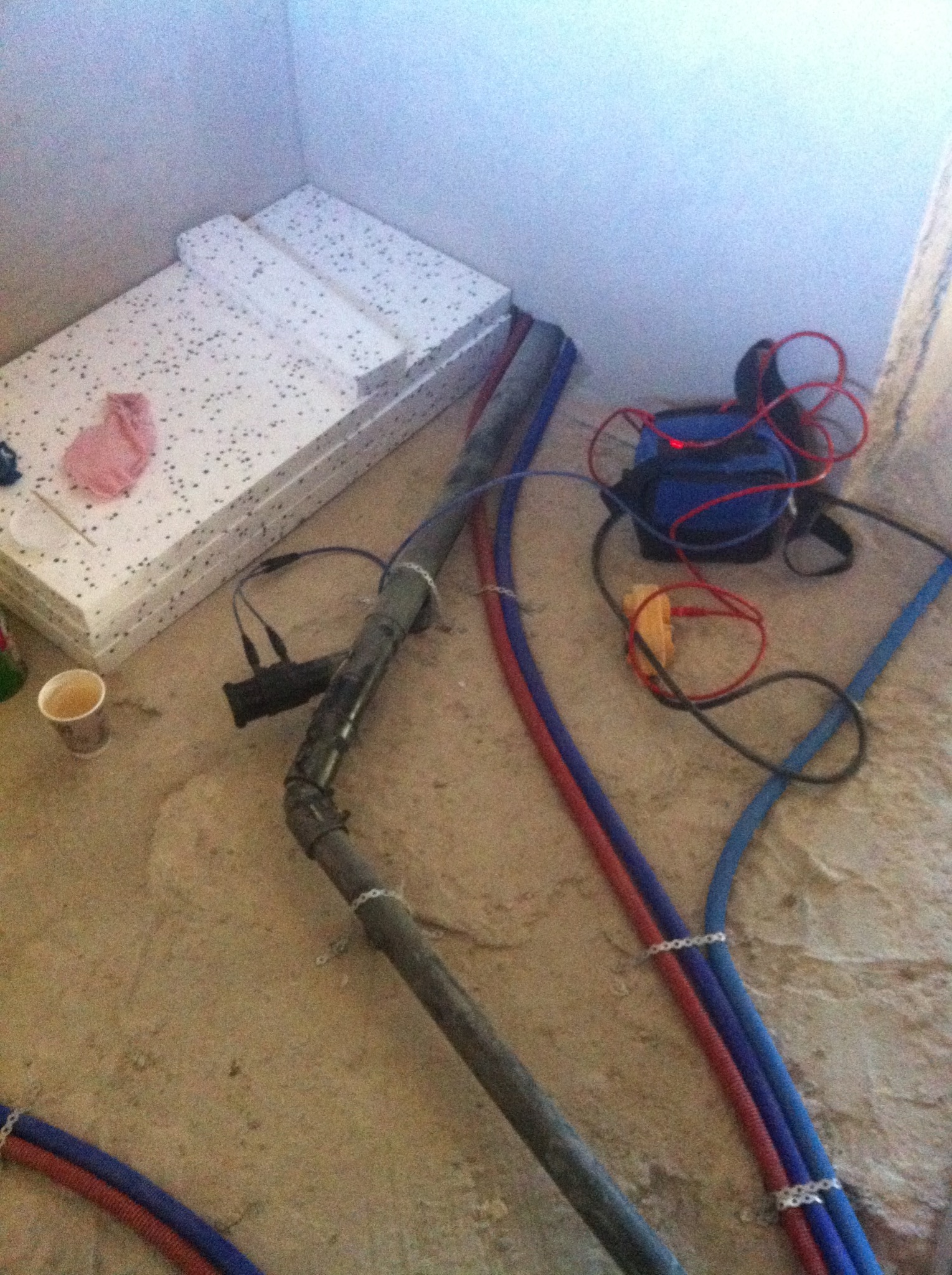 Przygotowania do instalacji ogrzewania podłogowego: styropianowe płyty izolacyjne, rury grzewcze spięte klamrami, narzędzia oraz kubek po kawie na surowej posadzce.