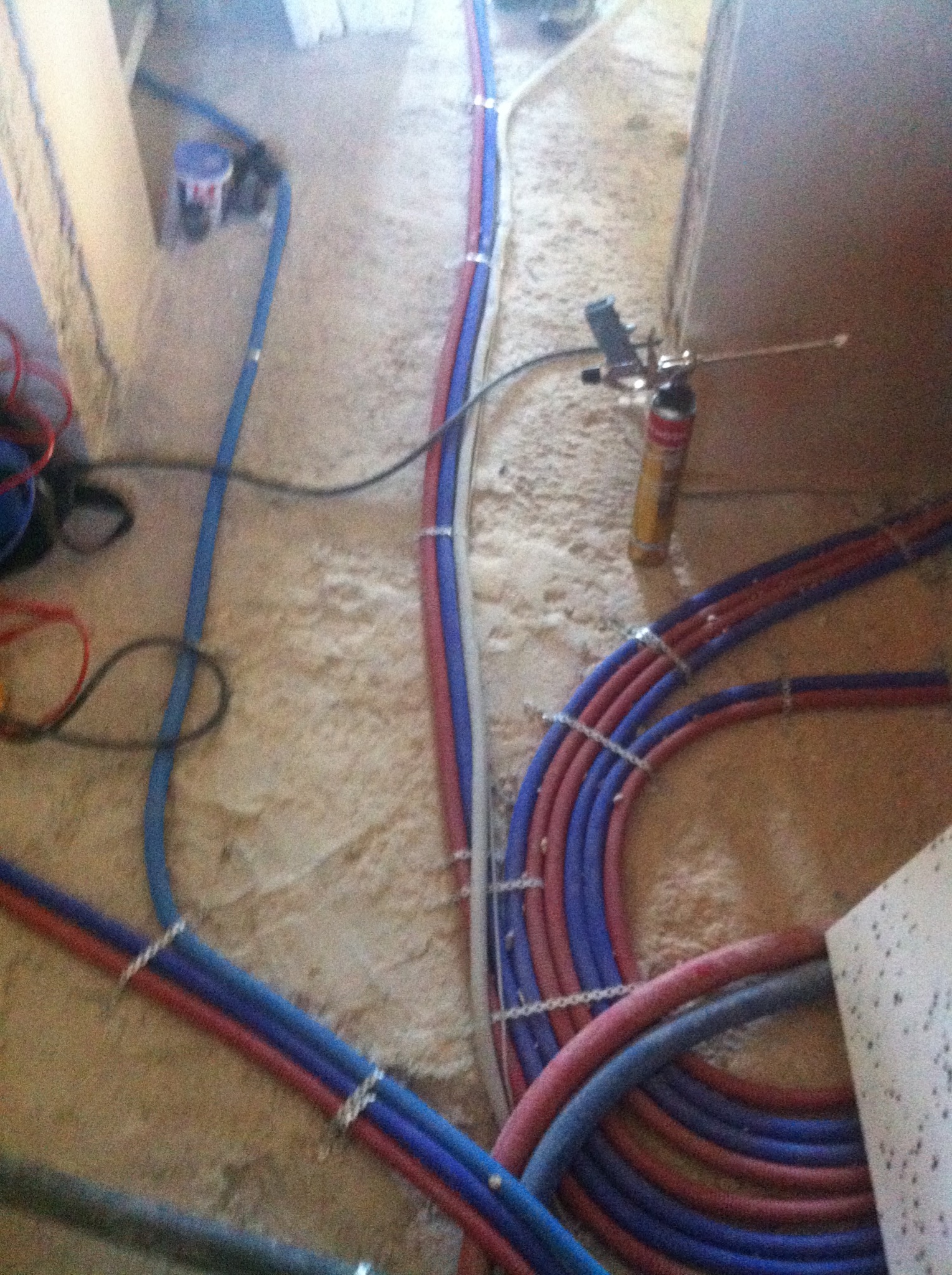 Układanie rur instalacji podłogowej, czerwone i niebieskie rury spięte klamrami na surowym podłożu, widoczny pistolet do pianki montażowej i wiadro z zaprawą.