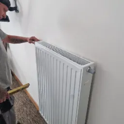 Instalator poziomuje biały grzejnik panelowy na ścianie, widoczny poziomica i fragment instalacji TV na ścianie. Ujęcie z boku, z bliska.