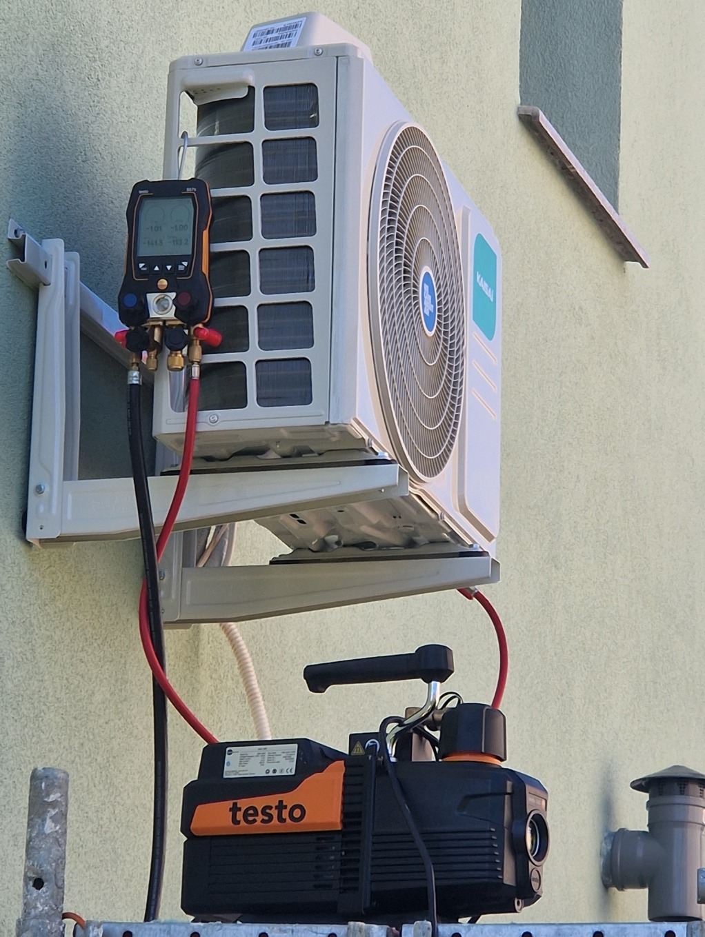 Biała jednostka zewnętrzna klimatyzacji marki Kaisai zamontowana na ścianie, podłączona do pompy próżniowej Testo i cyfrowego kolektora. Widoczne przewody i elementy montażowe.