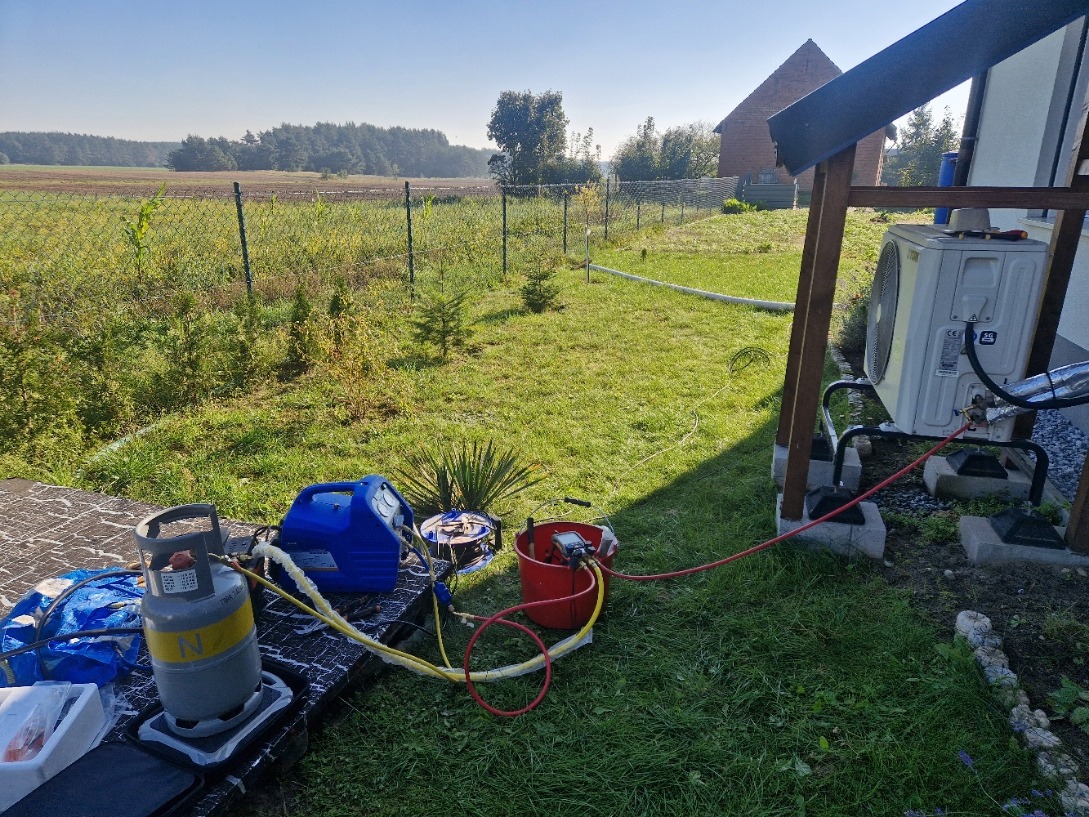 Montaż klimatyzacji zewnętrznej: jednostka przy budynku, narzędzia na stole w ogrodzie, wiejska sceneria w tle. Widoczne przewody i butla z gazem.