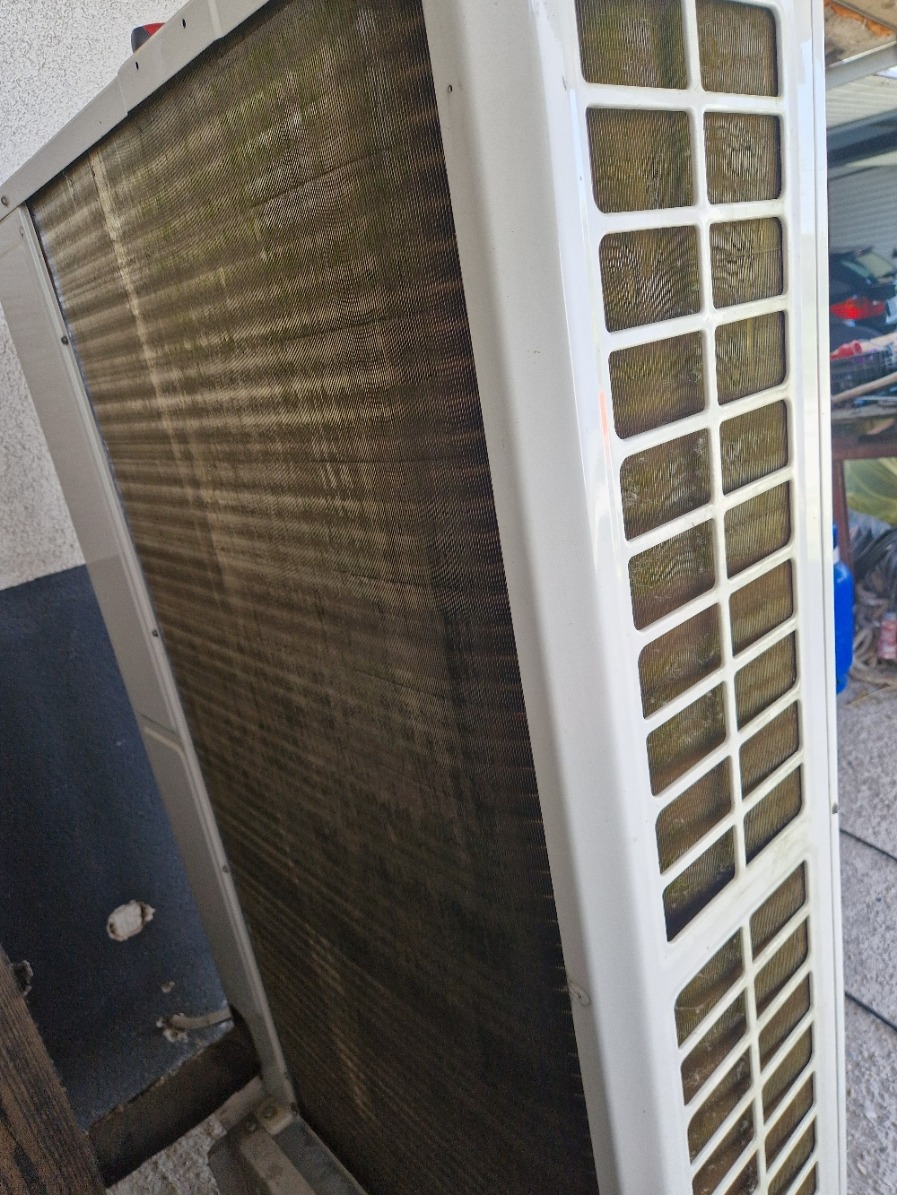 Zanieczyszczony wymiennik ciepła klimatyzatora z widocznymi lamelami i obudową z kratką wentylacyjną. Widoczne ślady zabrudzeń i zużycia.