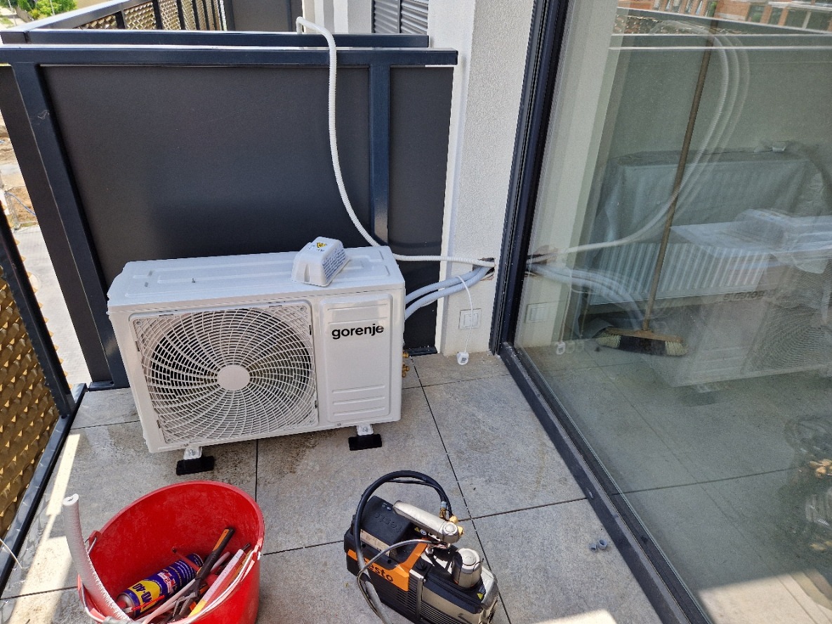 Biała klimatyzacja Gorenje na balkonie, z widocznym podłączeniem do ściany. Obok czerwone wiadro z narzędziami i pompa próżniowa.