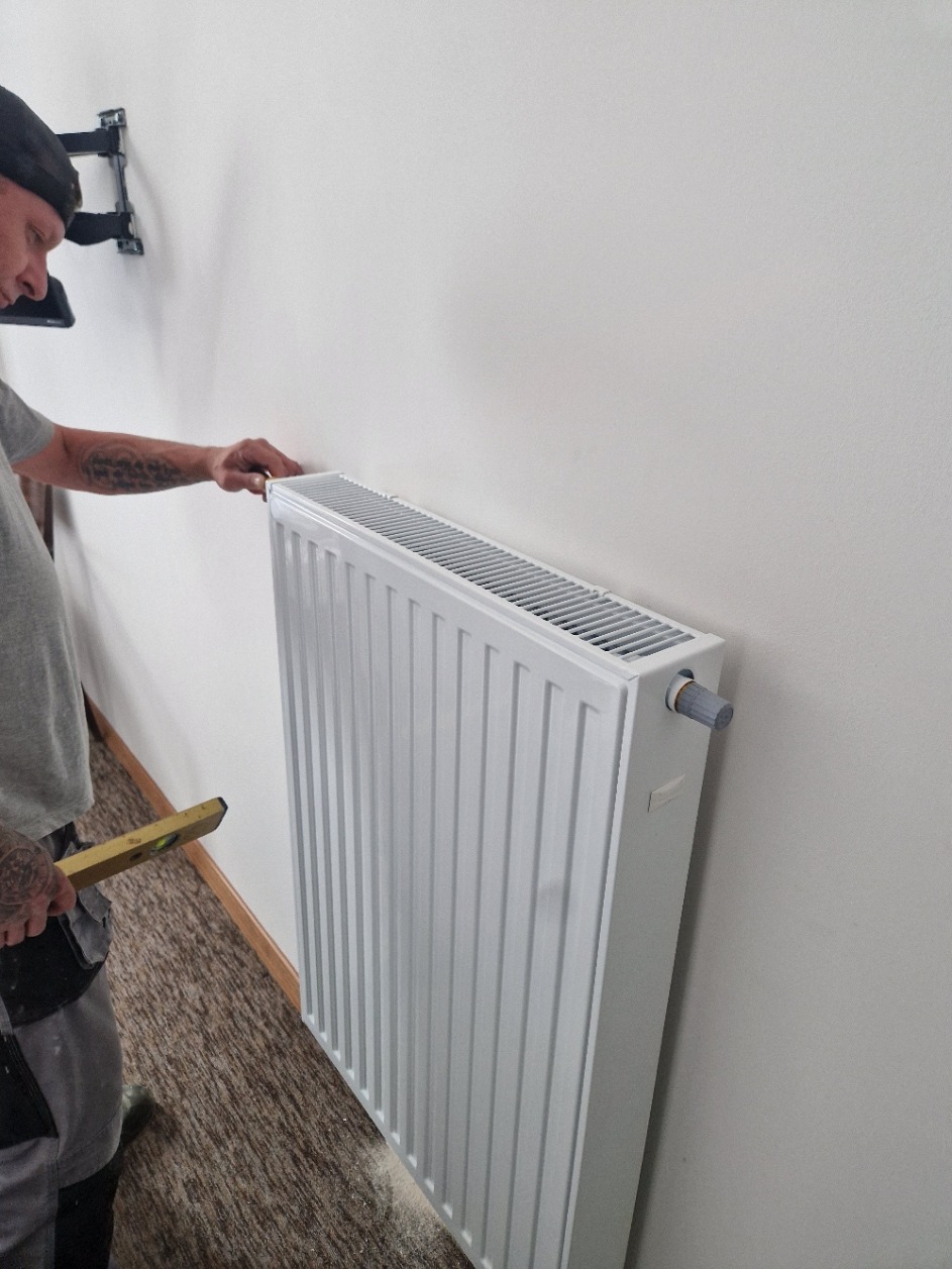 Instalator poziomuje biały grzejnik panelowy na ścianie, widoczny poziomica i fragment instalacji TV na ścianie. Ujęcie z boku, z bliska.