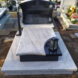 Granitowy nagrobek z czarnym elementem dekoracyjnym i lampionem, otoczony innymi nagrobkami na cmentarzu.