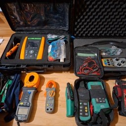 MP TEC - Zestaw profesjonalnych mierników elektrycznych różnych typów, w tym cęgowe, testery kabli i mierniki rezystancji, ułożonych na drewnianej podłodze w otwartych walizkach i torbach.