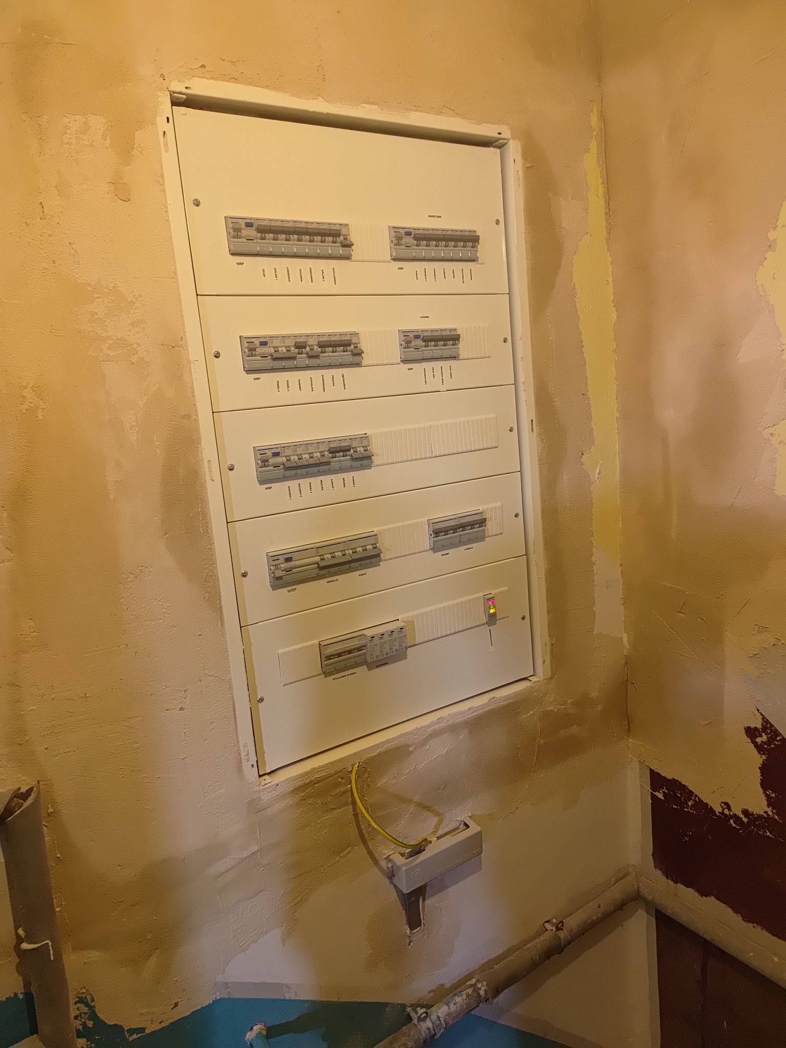 Rozdzielnica elektryczna z bezpiecznikami, zamontowana na nierównej, odrapanej ścianie w trakcie remontu, widoczny kabel uziemiający i rury.