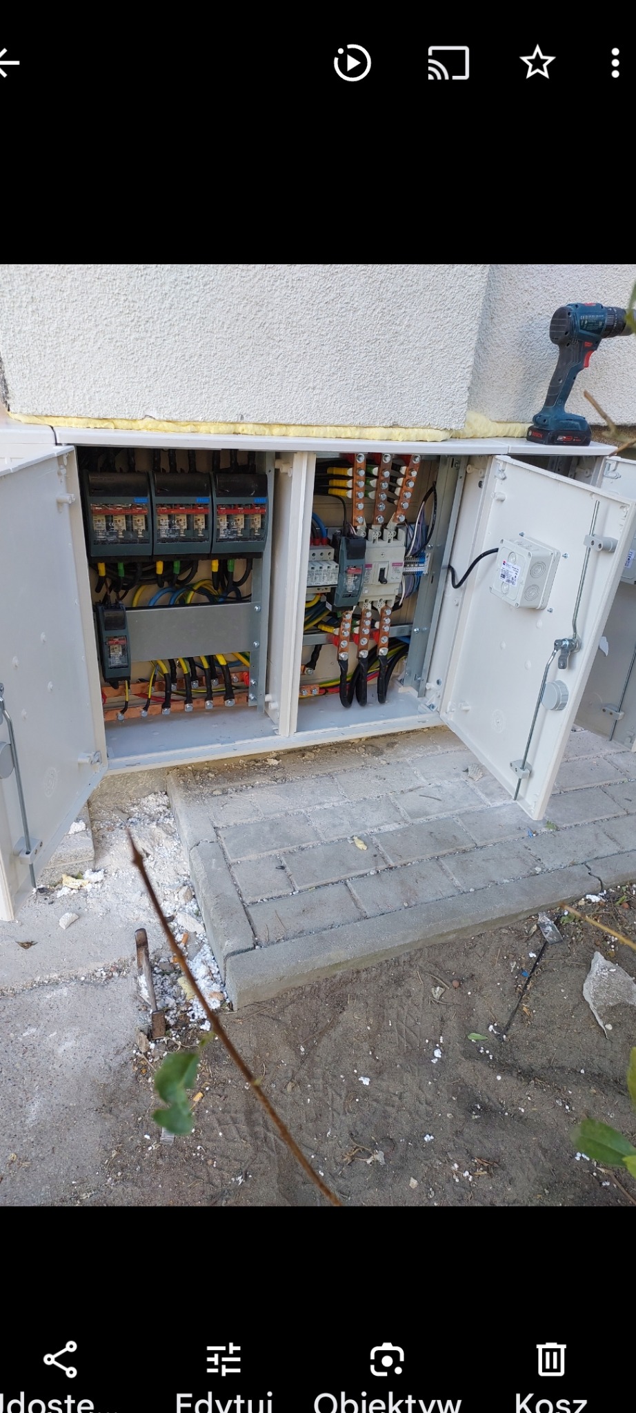 Otwarta skrzynka elektryczna zewnętrzna z widocznymi bezpiecznikami, przewodami i elementami instalacji, obok leży wkrętarka akumulatorowa.