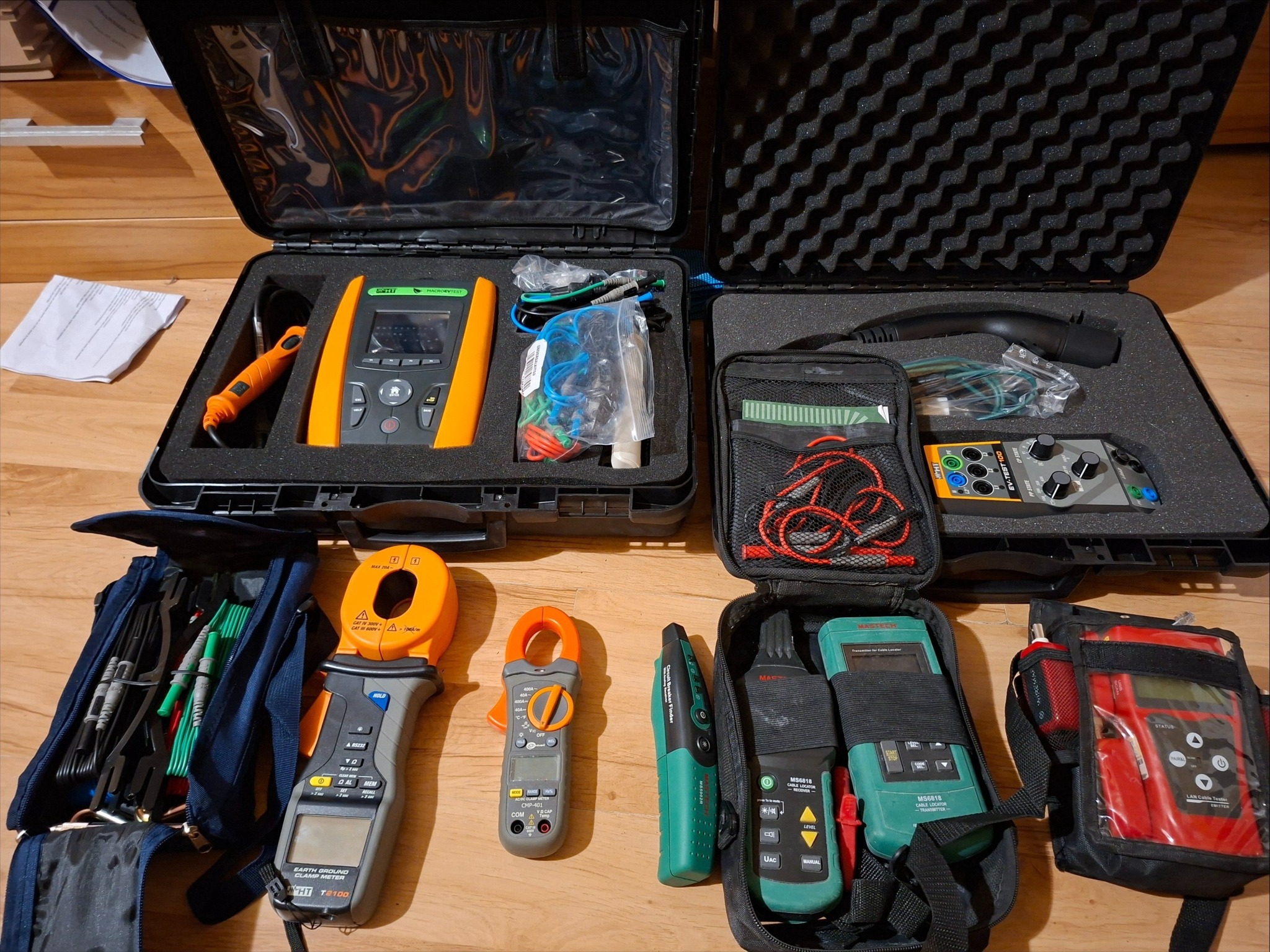 Zestaw profesjonalnych mierników elektrycznych różnych typów, w tym cęgowe, testery kabli i mierniki rezystancji, ułożonych na drewnianej podłodze w otwartych walizkach i torbach.