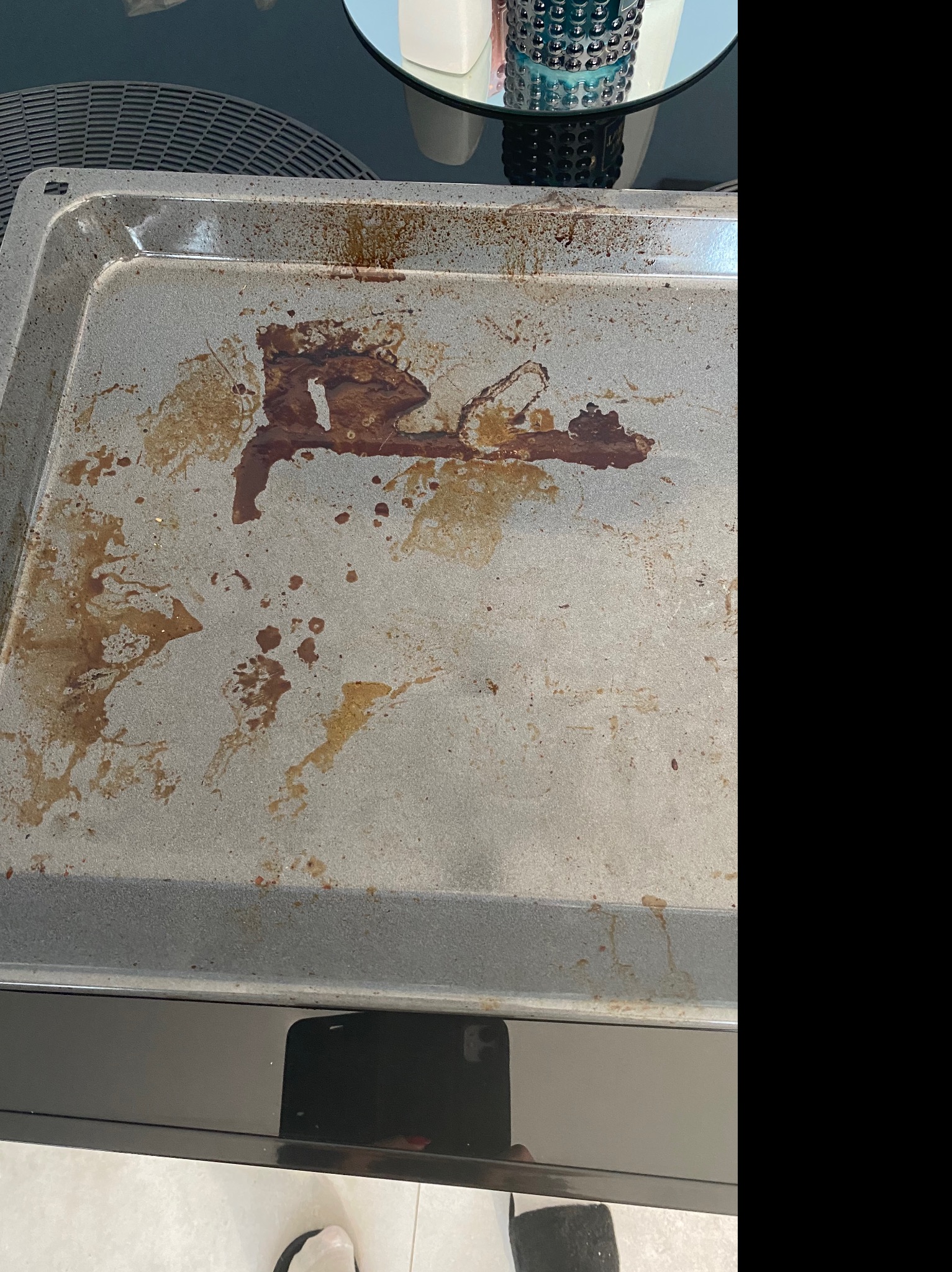 Brudna blacha do pieczenia z zaschniętymi plamami tłuszczu i resztek jedzenia, widoczna na tle czarnego piekarnika i fragmentu białej podłogi.