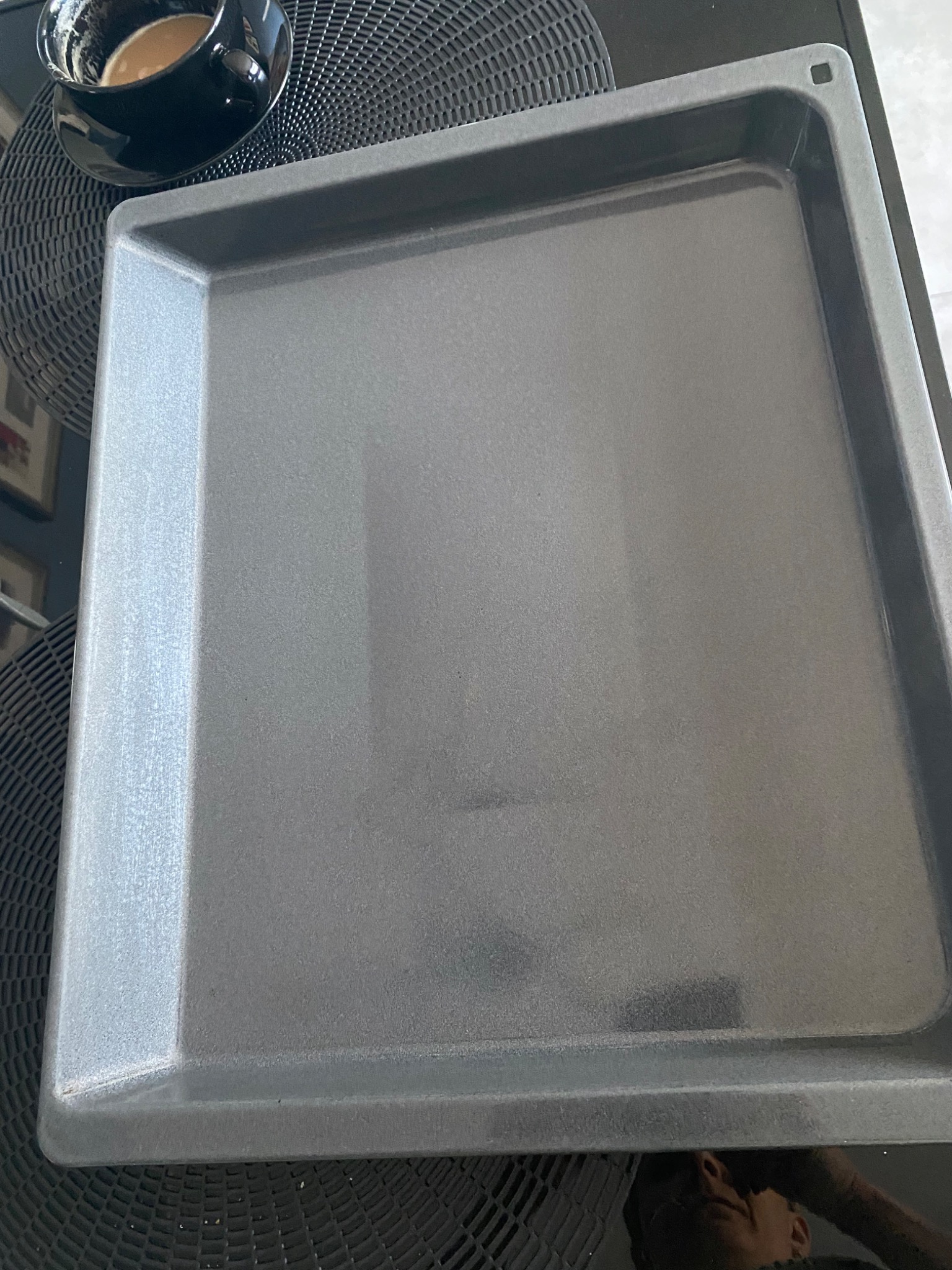 Szara, pusta blacha do pieczenia na czarnym, wzorzystym blacie, obok filiżanka kawy z mlekiem, ujęcie z góry.