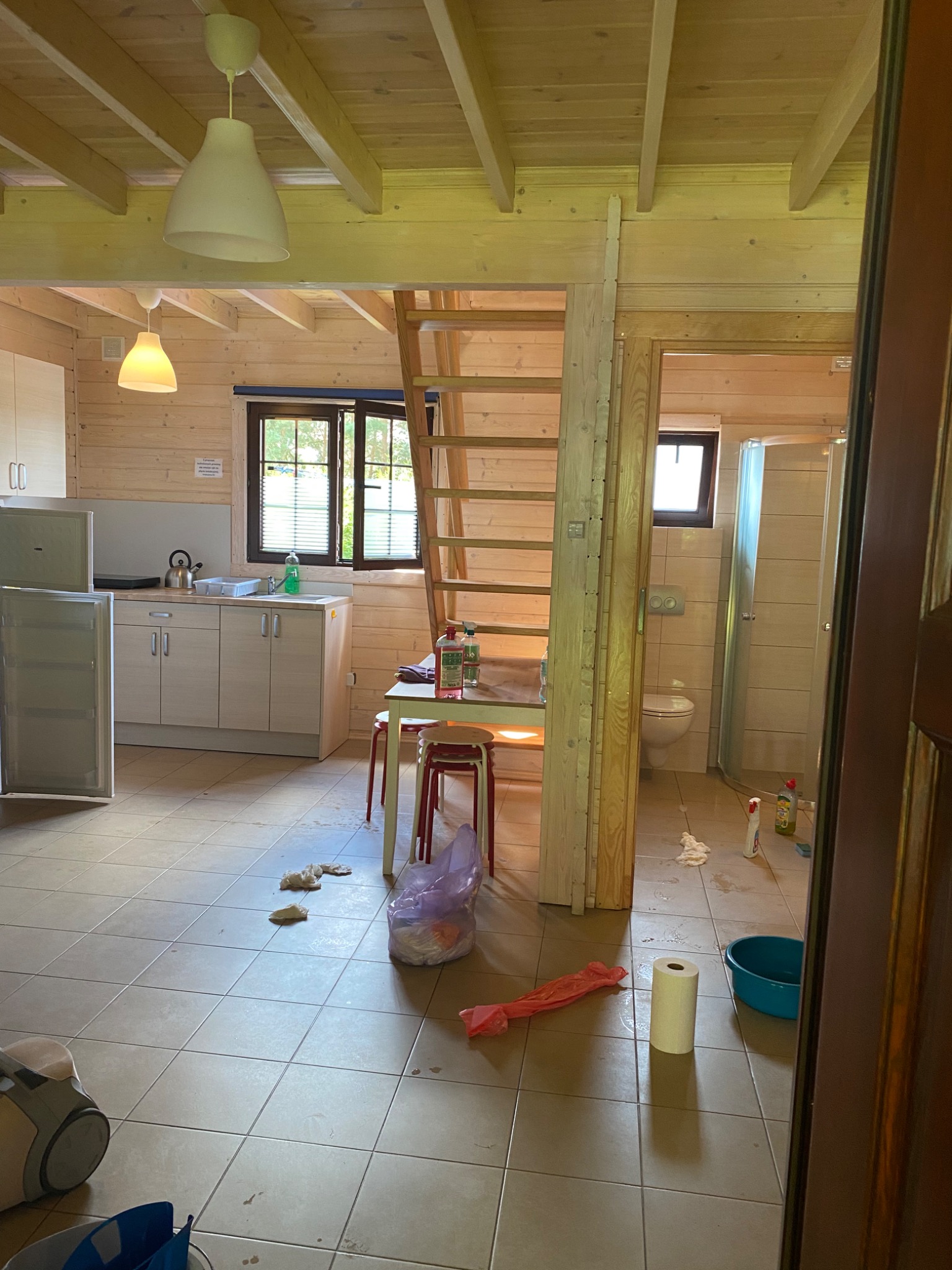 Wnętrze drewnianego domku przed sprzątaniem, widoczne ślady po remoncie, niedbale rozłożone przedmioty, drabina prowadząca na antresolę i otwarte drzwi do łazienki.