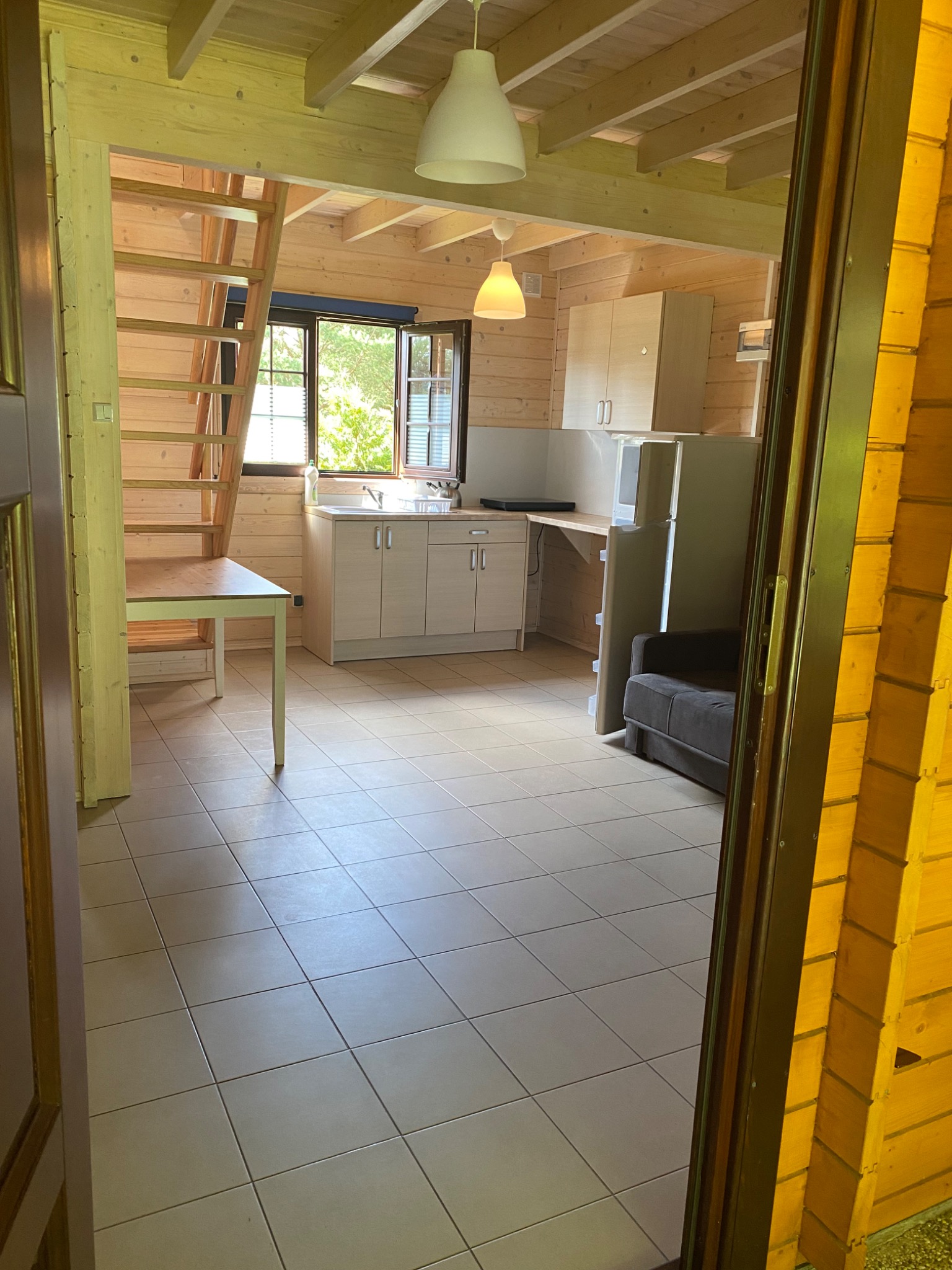 Wnętrze drewnianego domku letniskowego z aneksem kuchennym, schodami na antresolę i częścią wypoczynkową, widoczne z perspektywy wejścia.